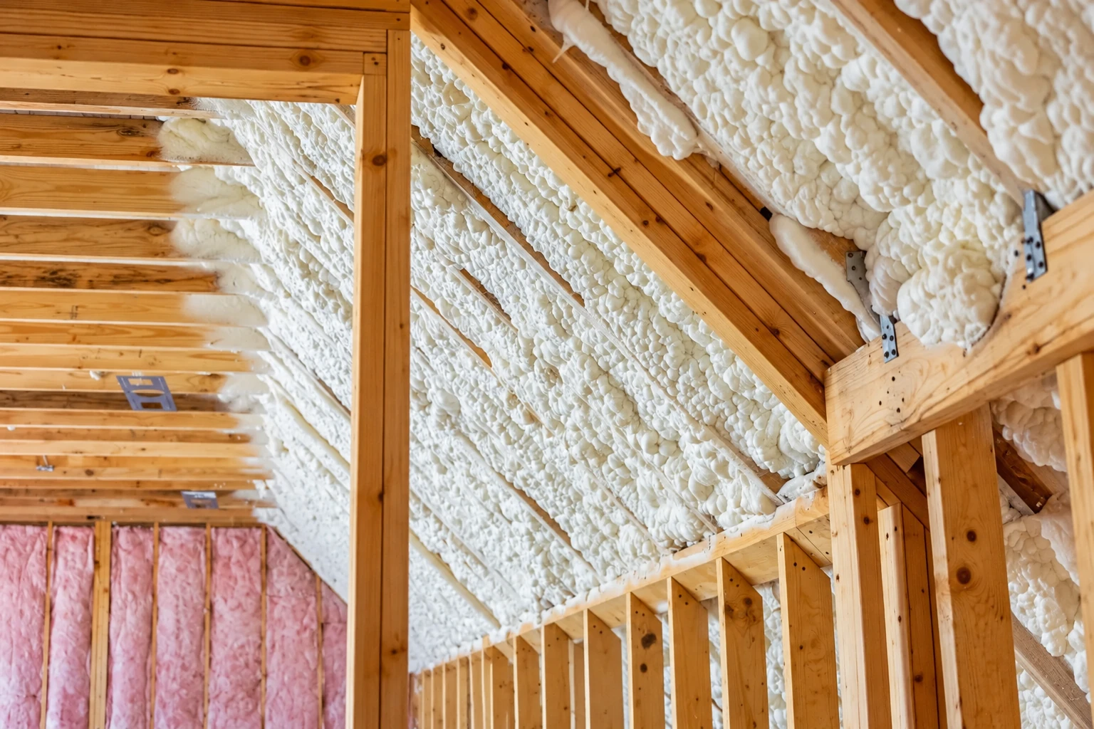 Exposed wooden roof framing packed with white spray foam insulation, with pink batt insulation visible in a lower corner.