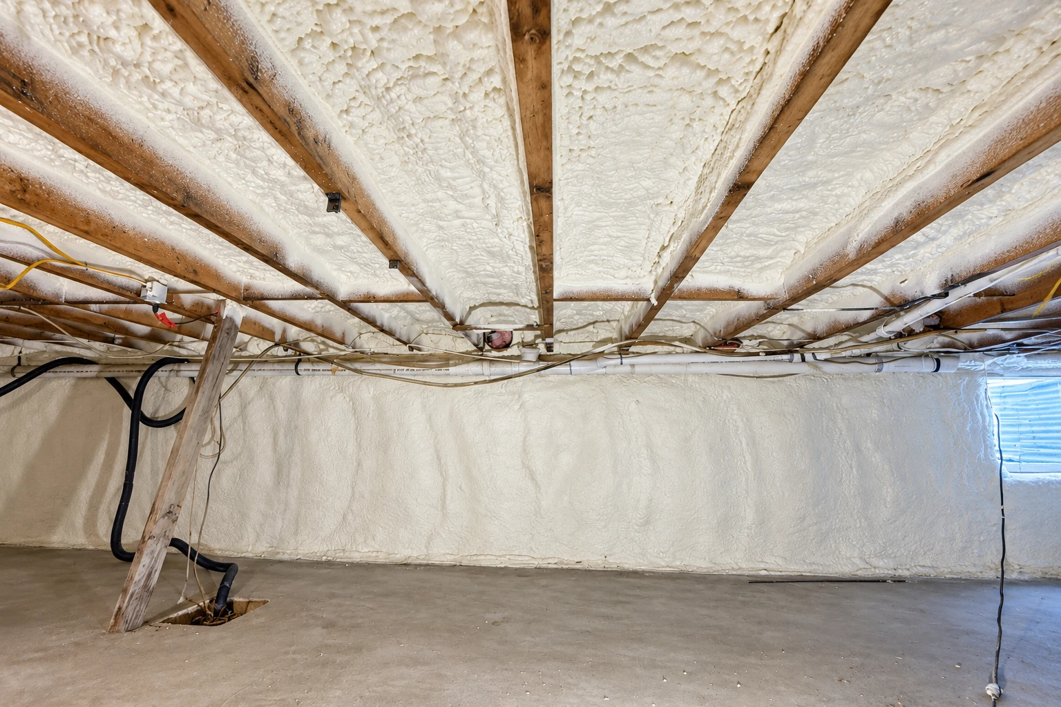 White spray foam coats the ceiling and walls of a crawl space, with exposed pipes and wires running across the ceiling.