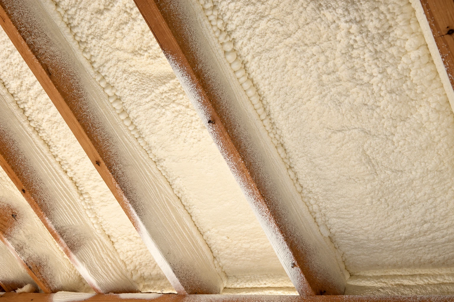 Cream spray foam insulation fills the spaces between exposed wooden rafters along an attic roof.