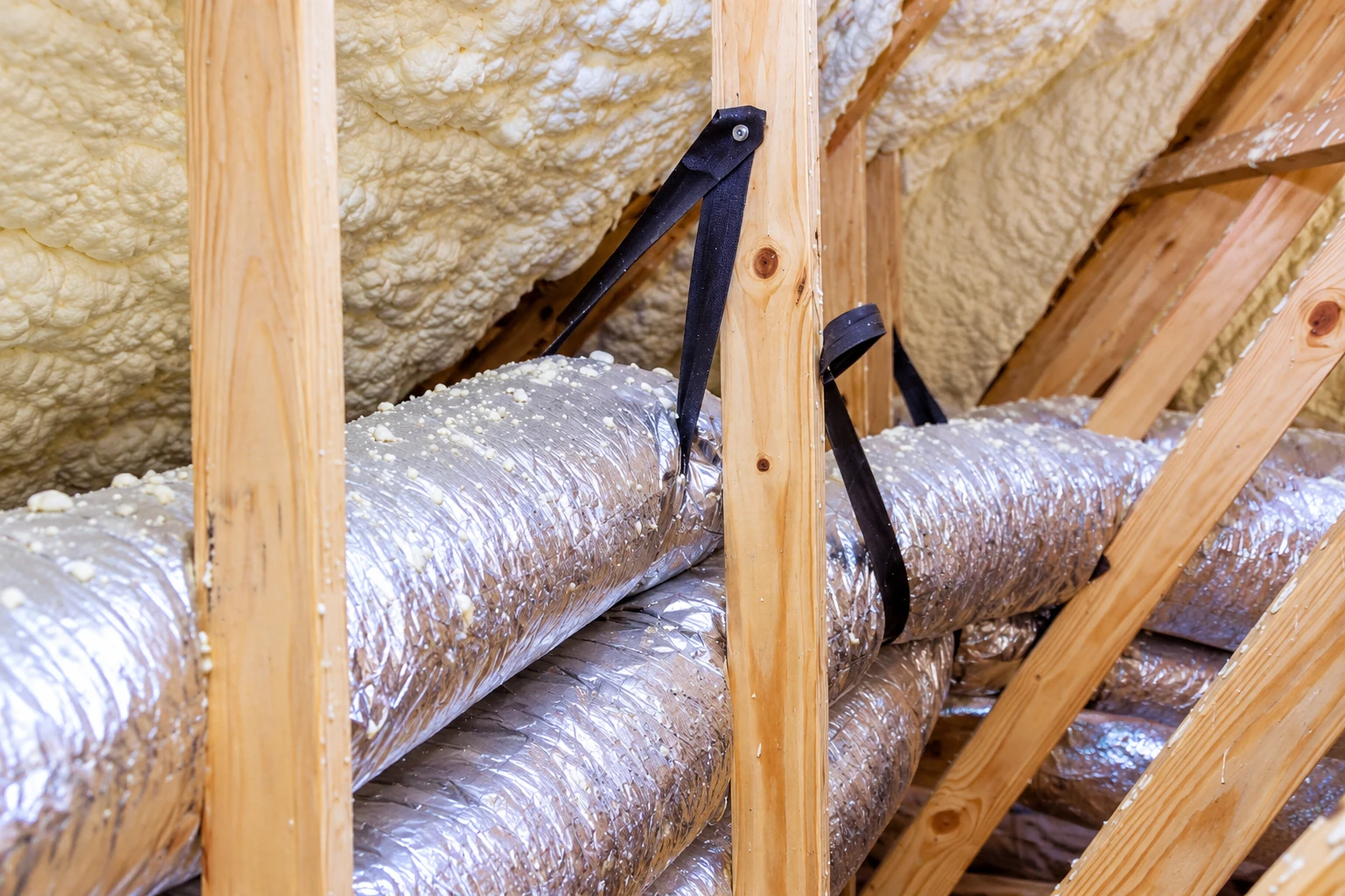 Foil-wrapped attic ducts run between wooden rafters beneath a roof coated in cream spray foam insulation.